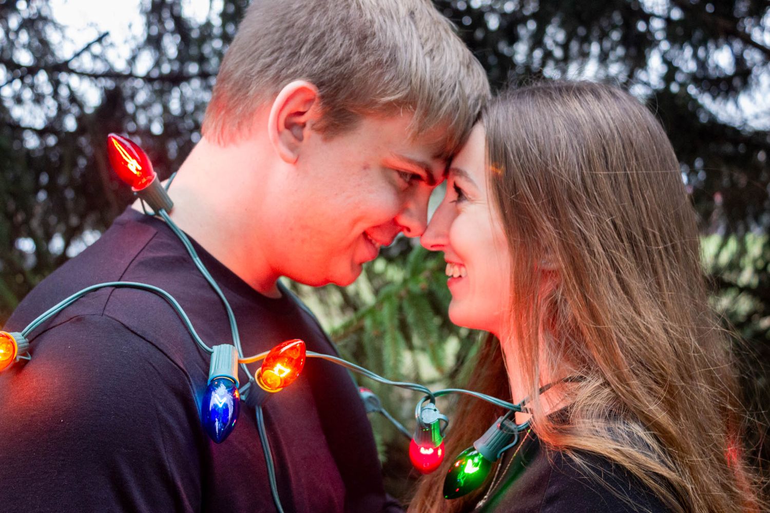 Romantic couple touching foreheads wrapped in colorful Christmas lights outdoors smiling.