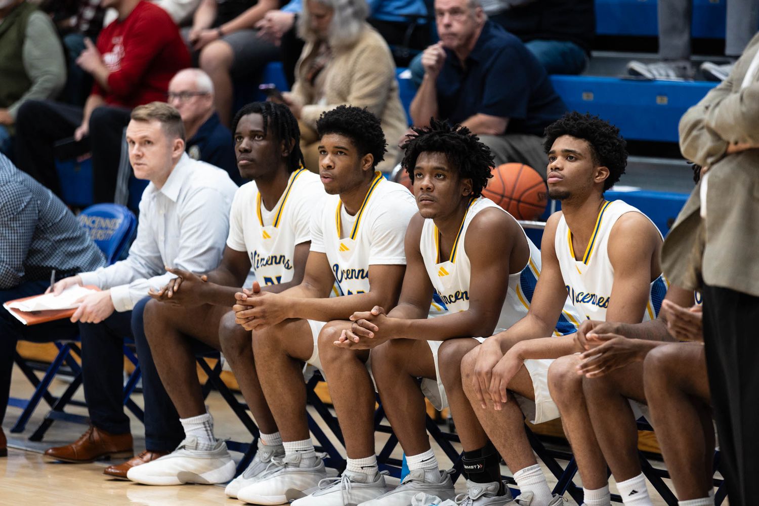 Vincennes University basketball players sit on the bench watching the game with a coach.