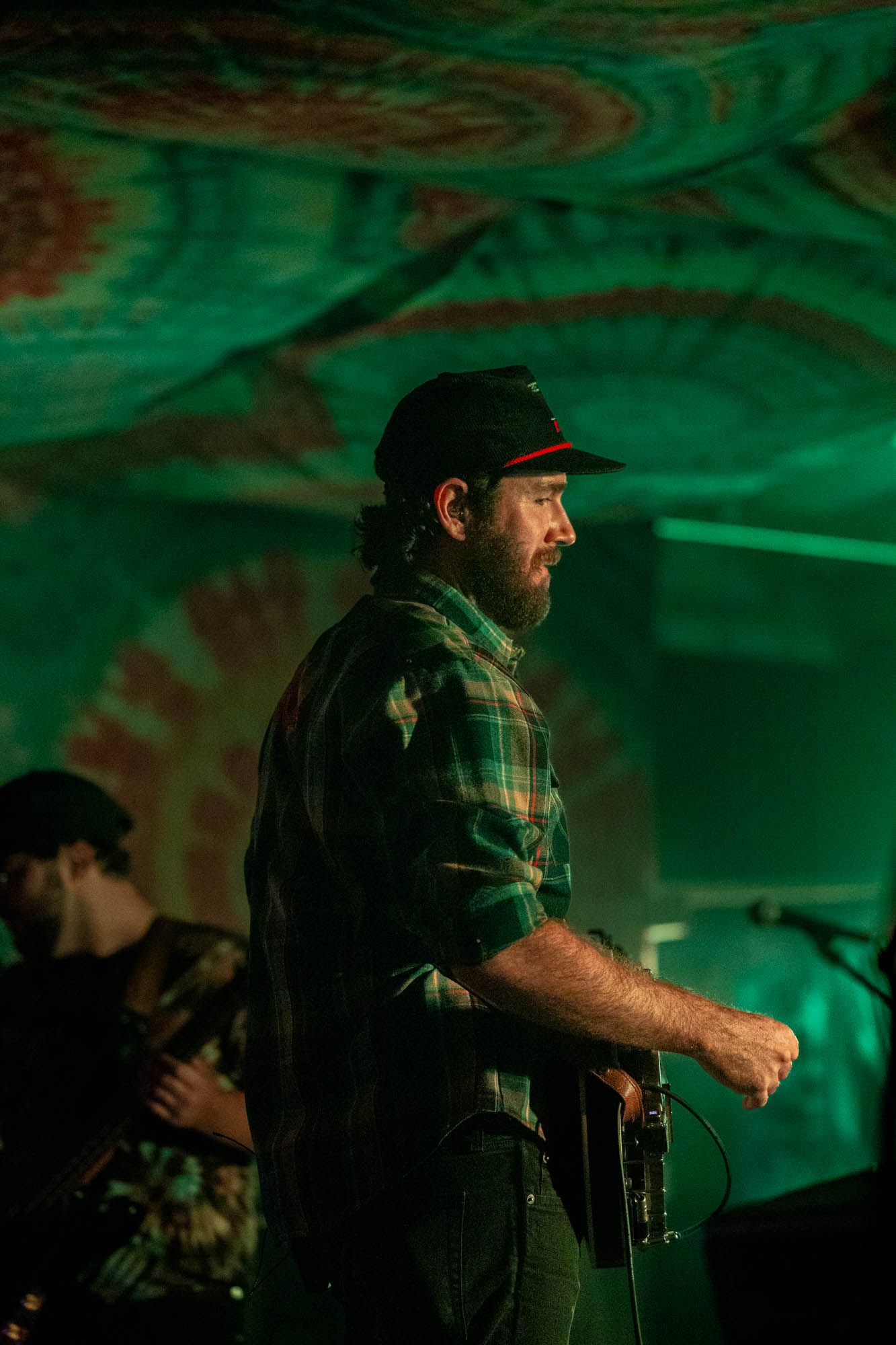 Bearded man in flannel shirt and cap plays instrument on dimly lit green-lit stage.
