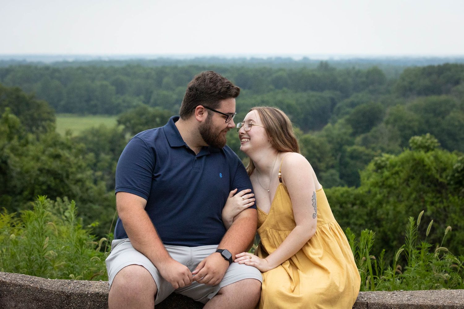 A couple sits on a stone wall smiling at each other with lush green trees and fields in the background.