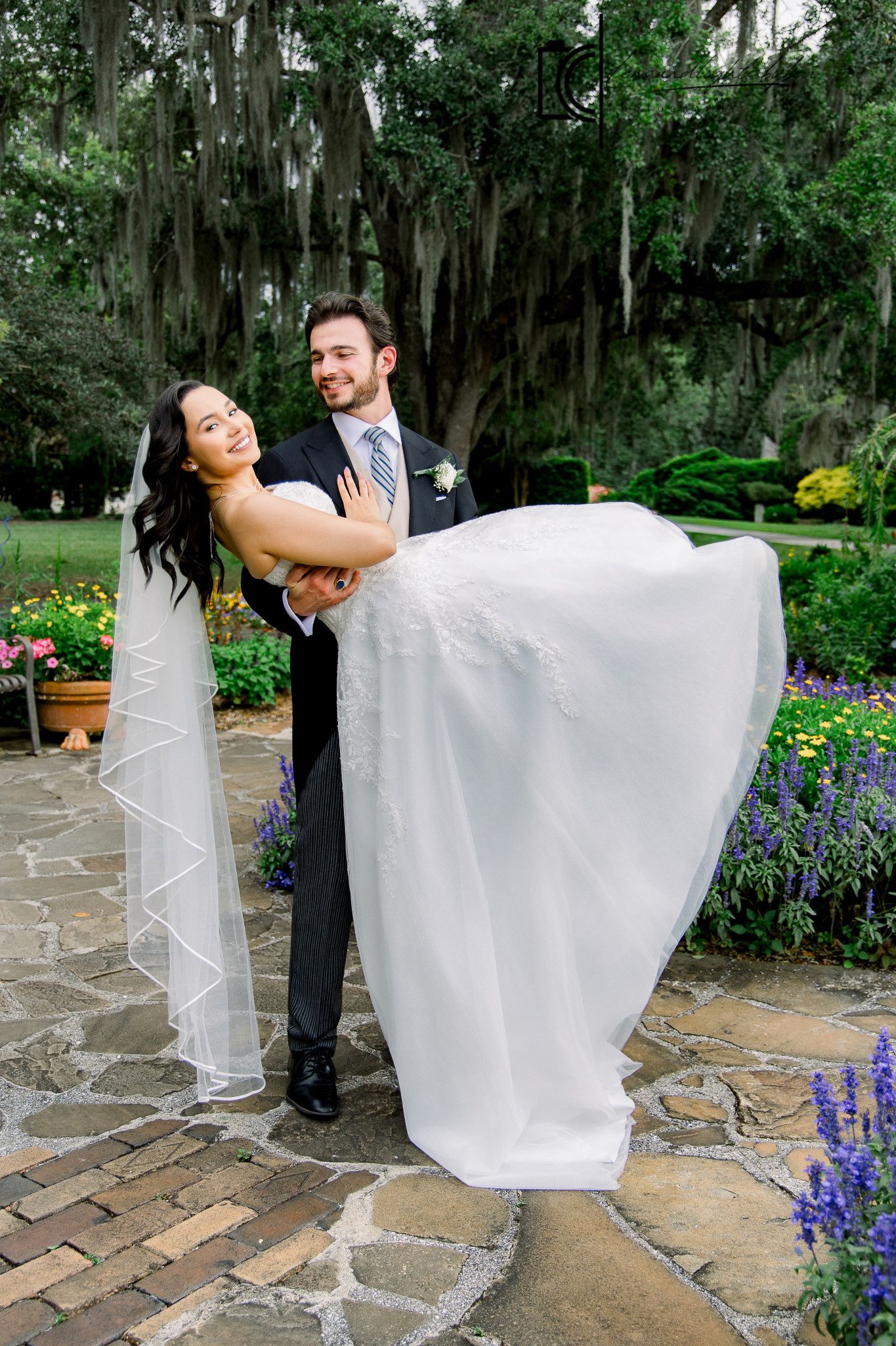Groom lifts smiling bride in white gown and veil outdoors on stone patio surrounded by lush garden greenery.