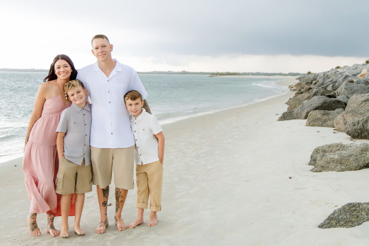 Family of four smiling together on a sandy beach with rocky jetty and calm ocean waves in the background.