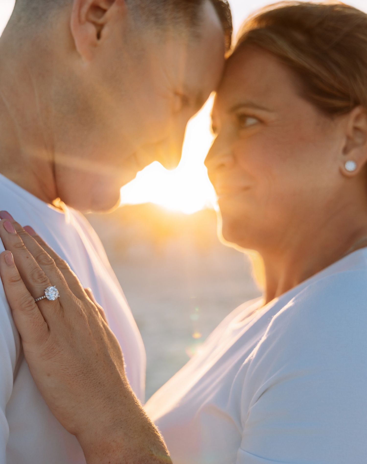Couple sharing a romantic sunset moment, woman wearing a diamond engagement ring, golden backlight glowing between them.