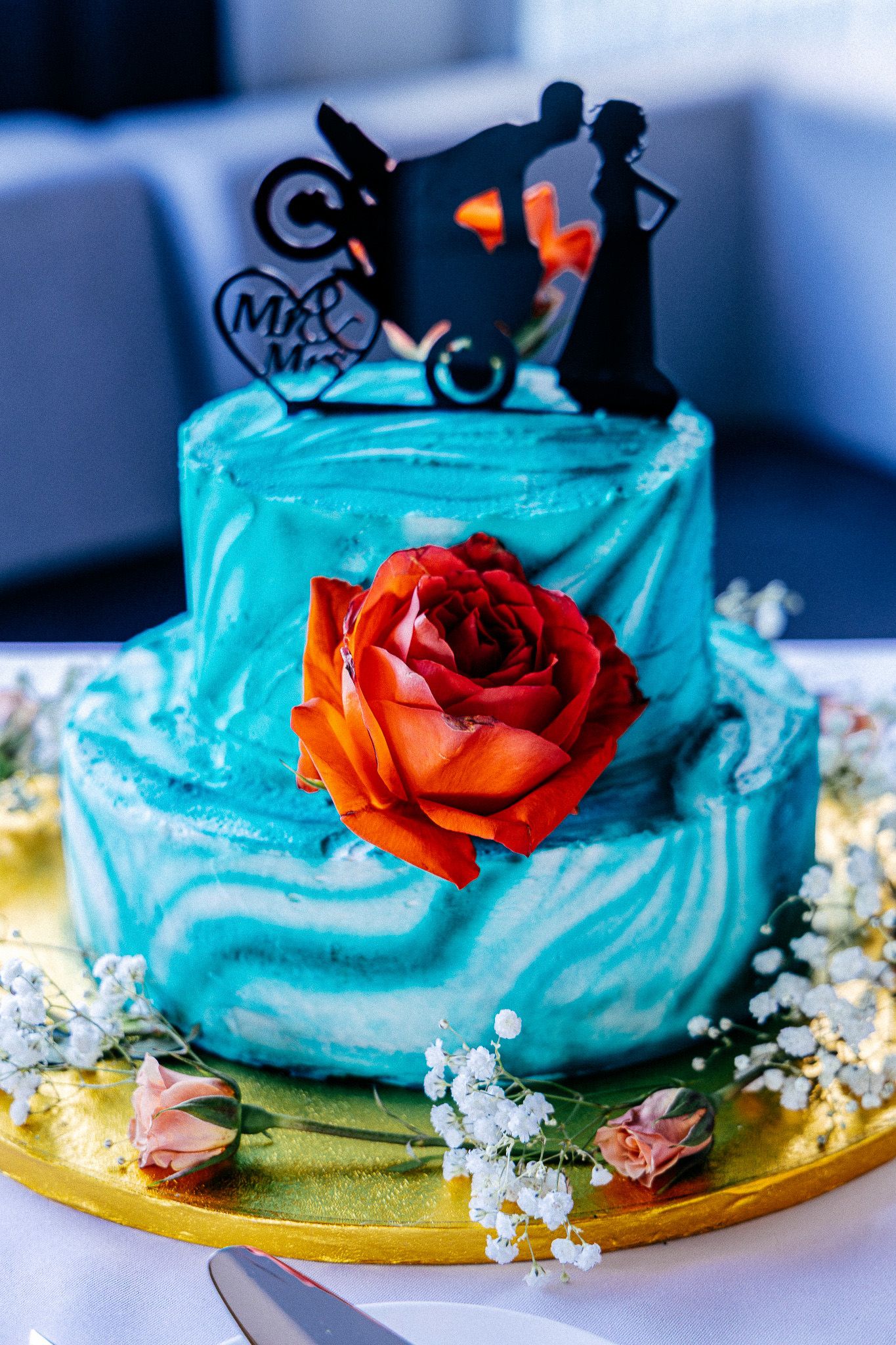 A turquoise blue cake decorated with an orange rose and motorcycle silhouette topper on a gold base with baby's breath flowers.