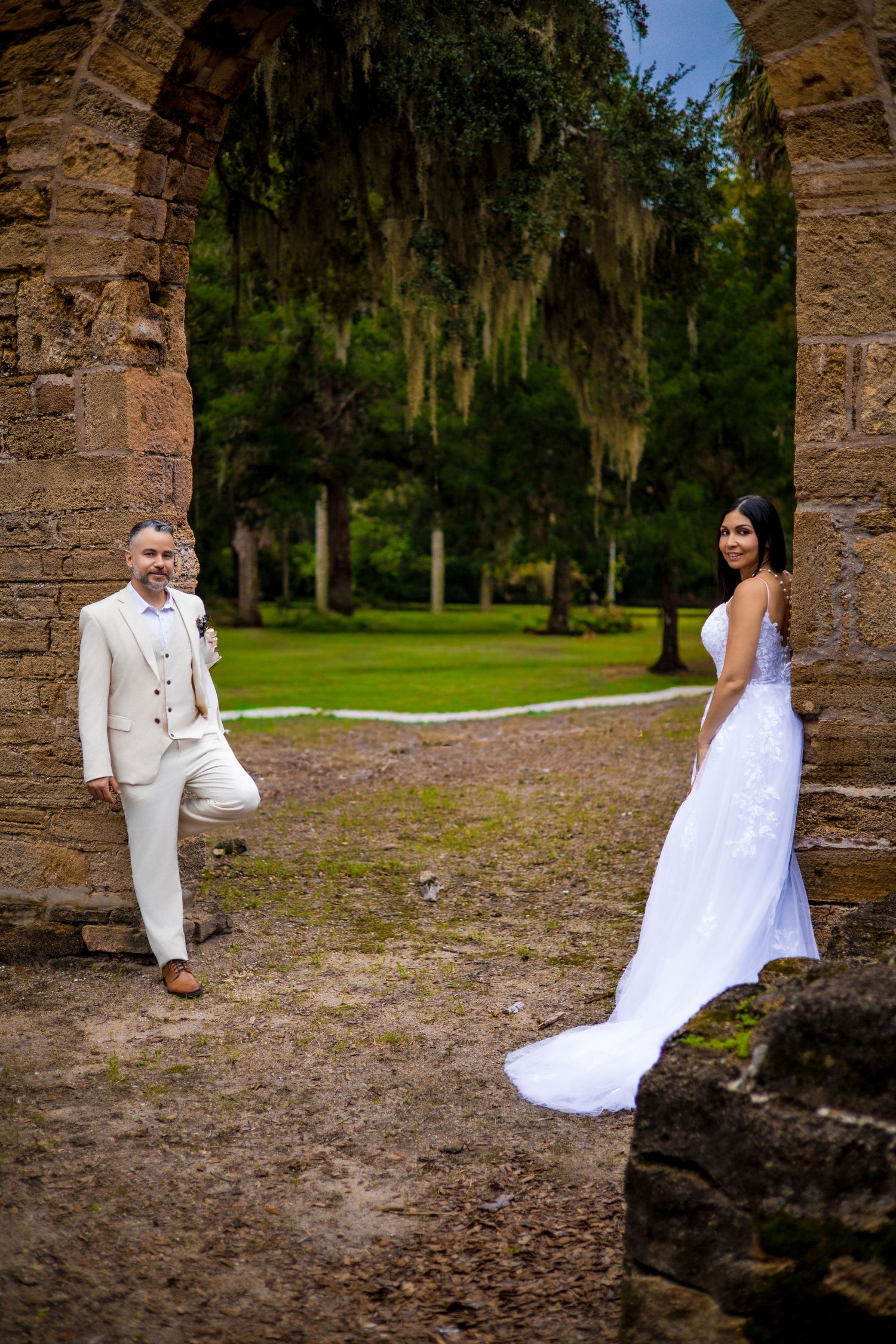 Wedding couple plays hide and seek around stone pillars in outdoor garden setting.