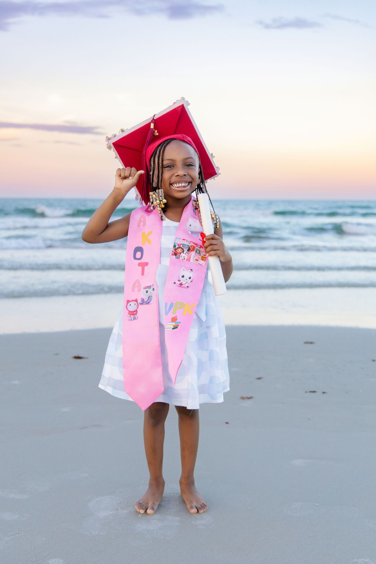 Young girl in red graduation cap and pink stole stands barefoot on beach at sunset, celebrating her achievement.