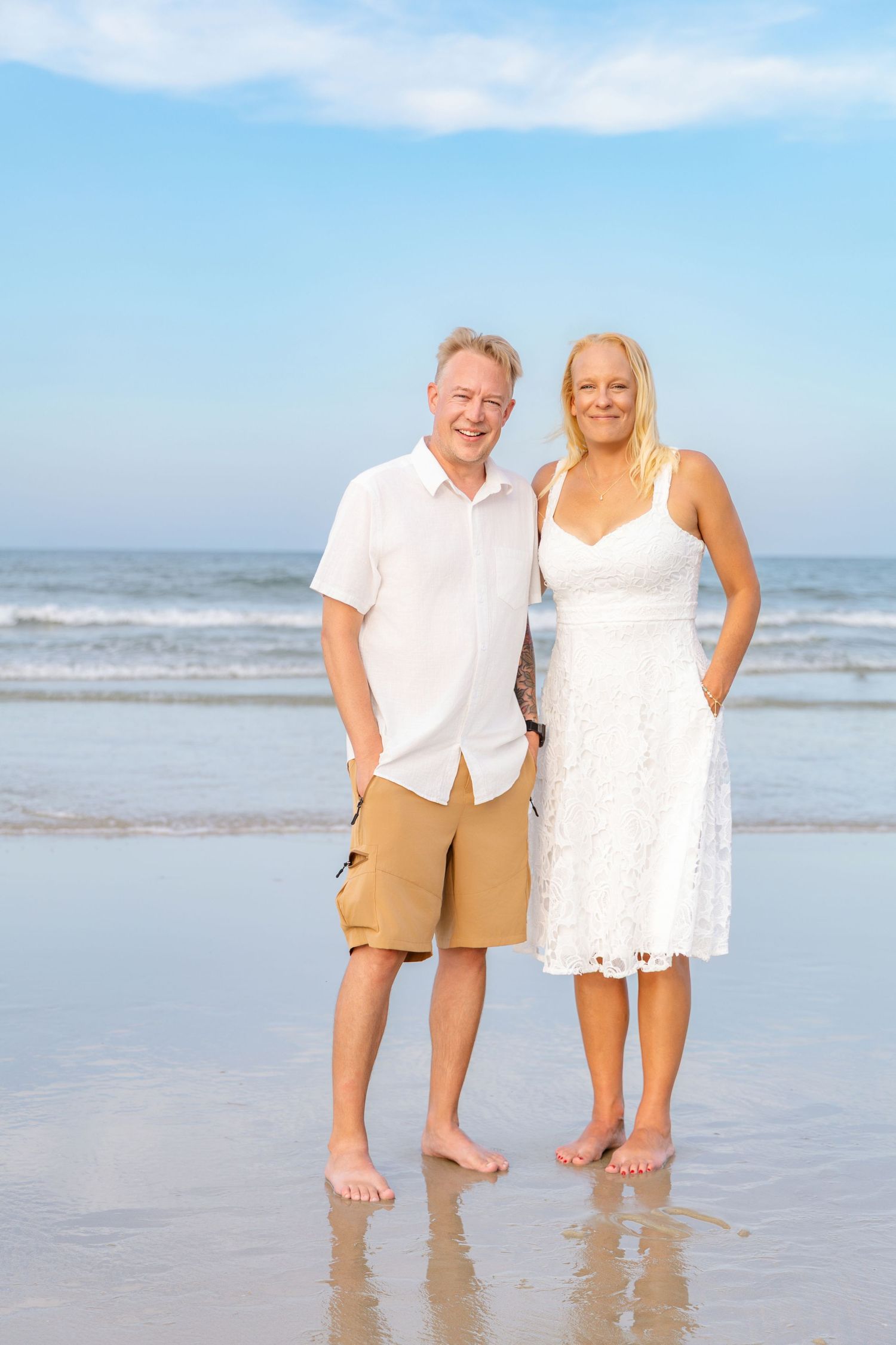 Happy mature couple standing barefoot on a sunny beach in white and tan outfits, smiling at camera.