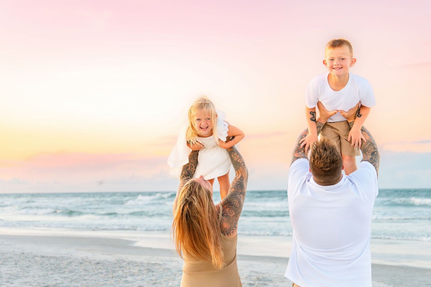 Family of four enjoying a beautiful sunset beach moment, with parents lifting their laughing children on their shoulders.