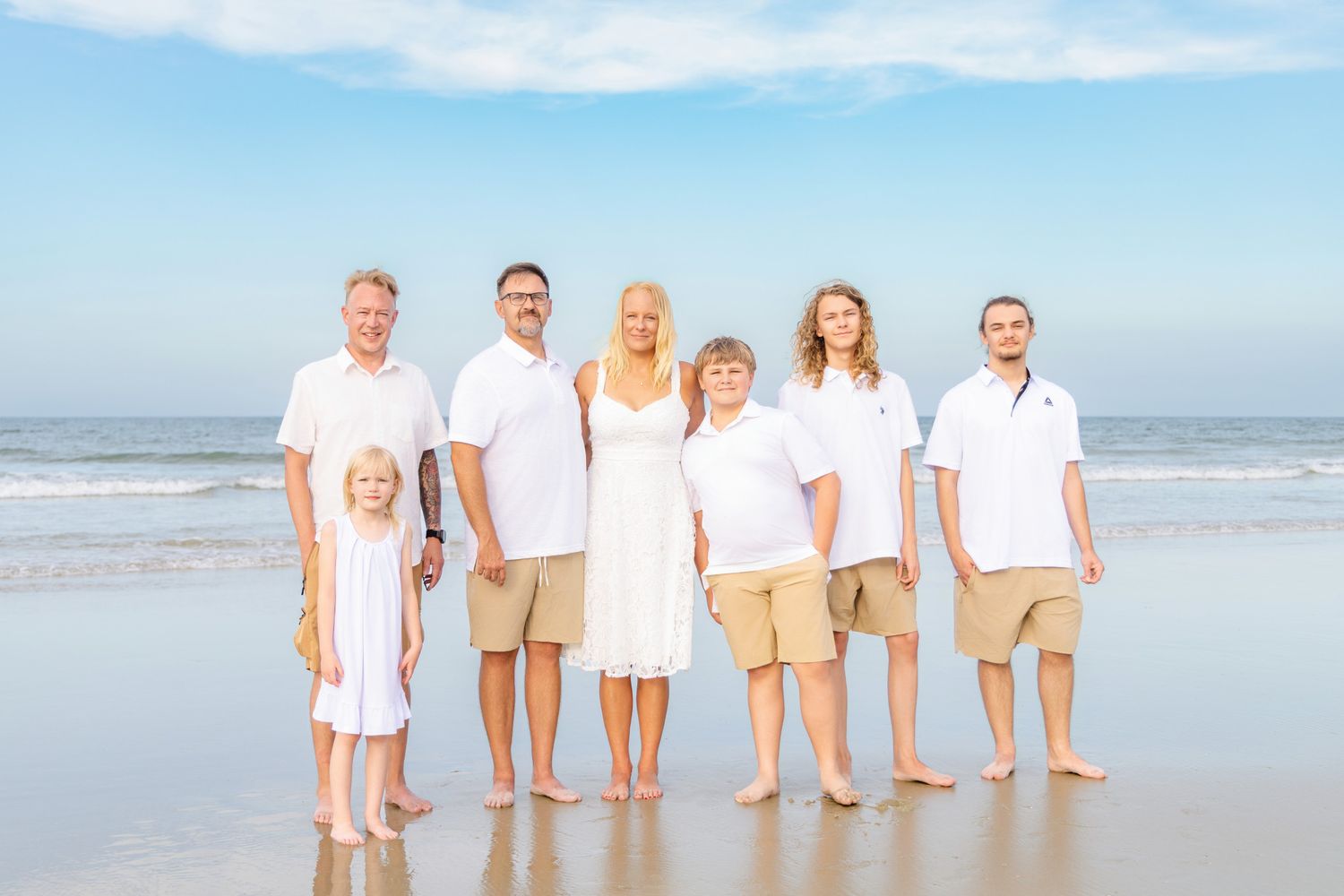 Family of seven posing together on a sunny beach, wearing white and khaki outfits, with ocean waves in the background.