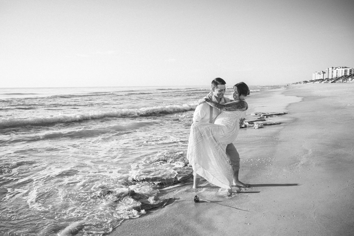 Black and white photo of newlyweds embracing playfully in ocean waves on sandy beach.