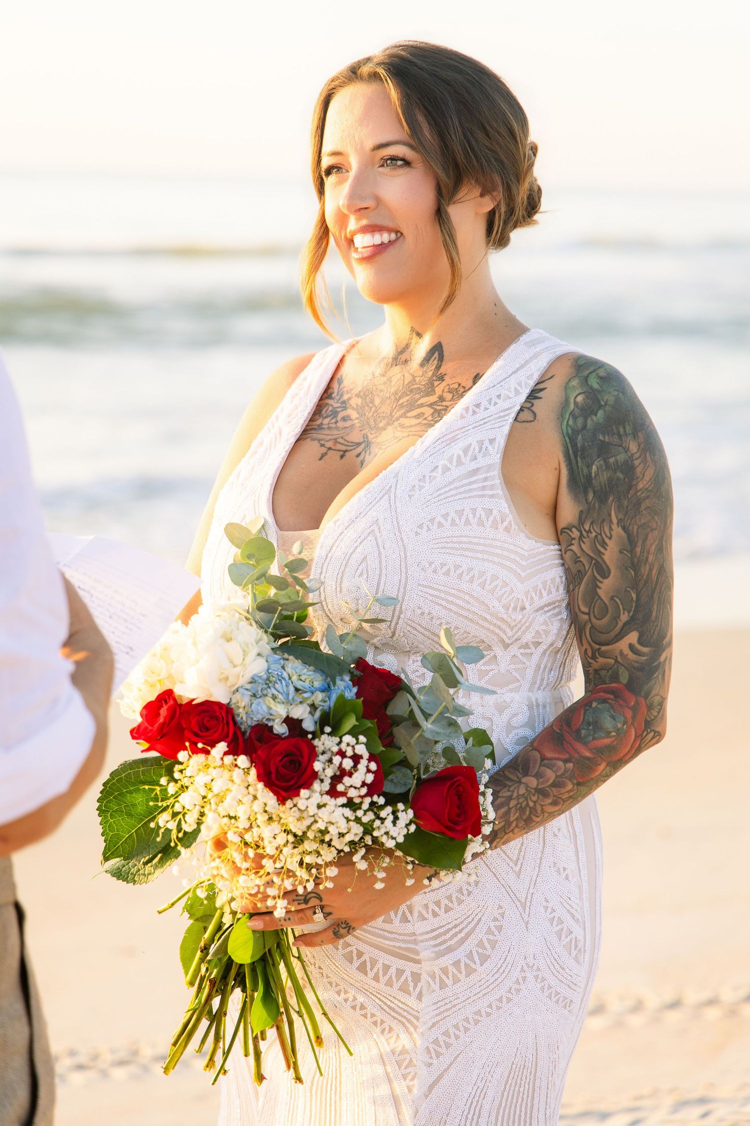 Radiant tattooed bride smiles holding red rose and hydrangea bouquet at beach wedding.
