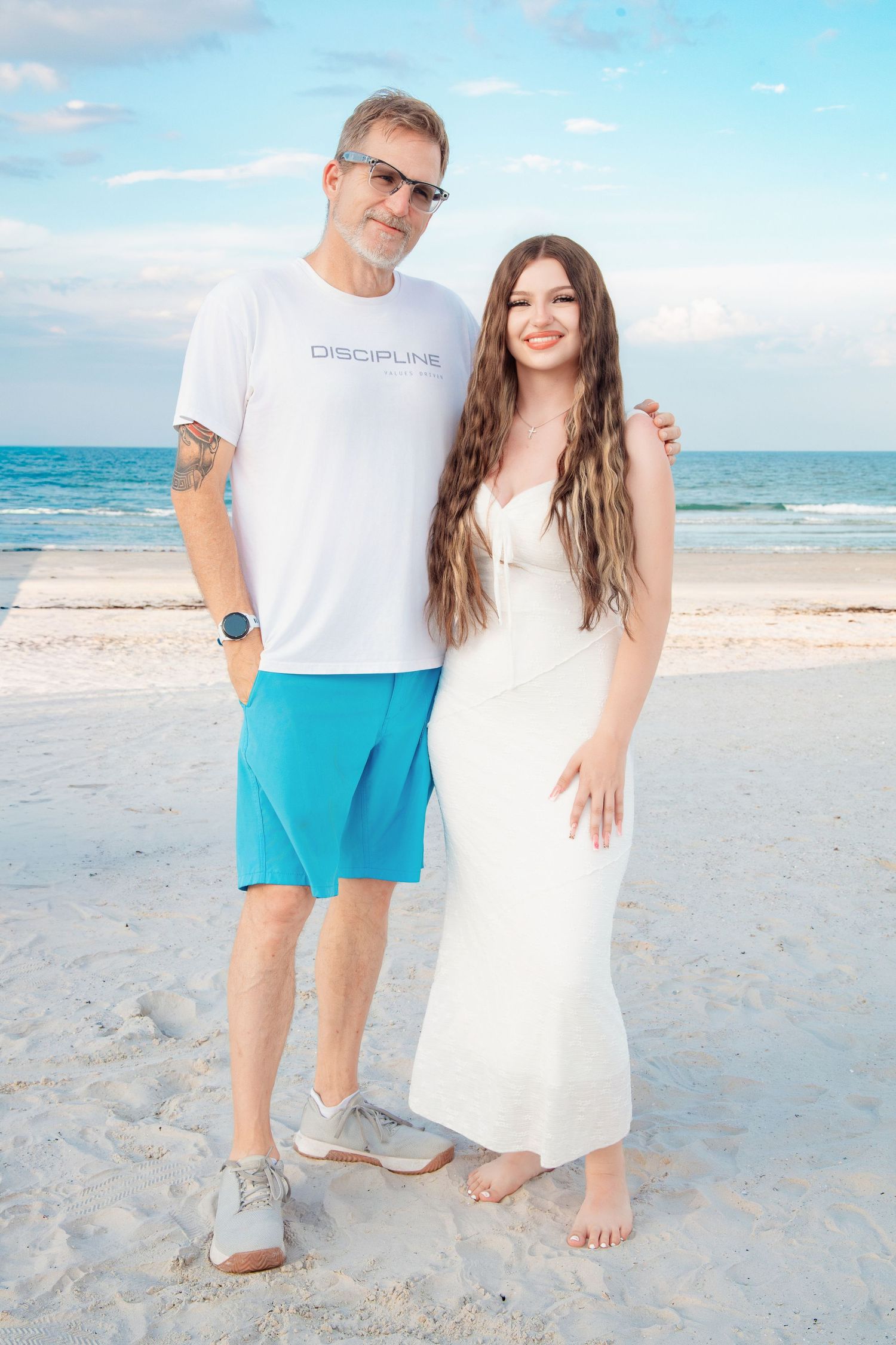 A man and young woman pose together on a sunny white sand beach, wearing white tops and turquoise shorts.