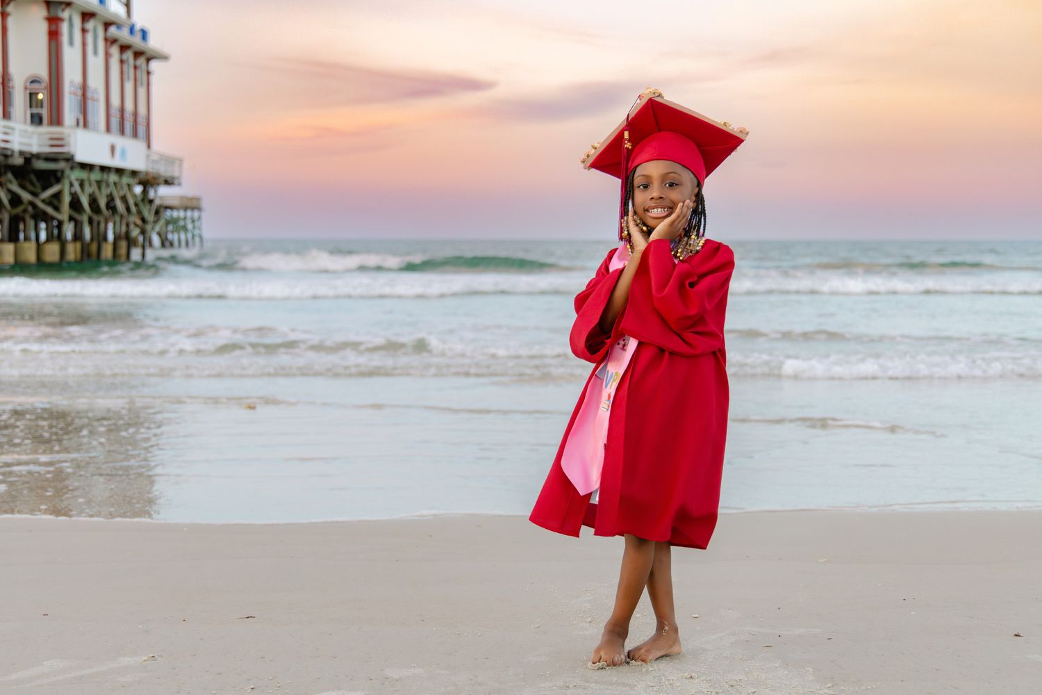 Young girl in red graduation cap and gown smiling on a beach at sunset with waves and pier in background.