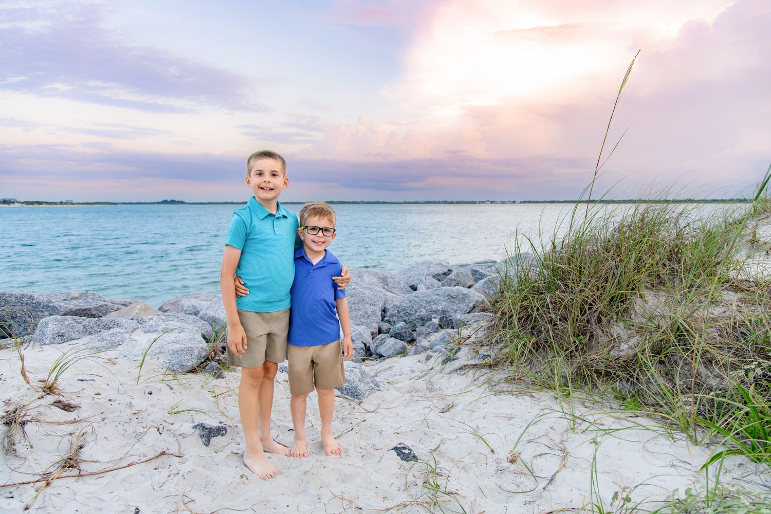 Two young boys stand smiling on a sandy beach at sunset, with turquoise water and rocks in the background.