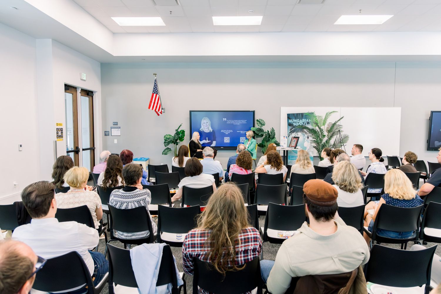 Audience members seated in rows attend a presentation in a modern conference room with an American flag and display screen.