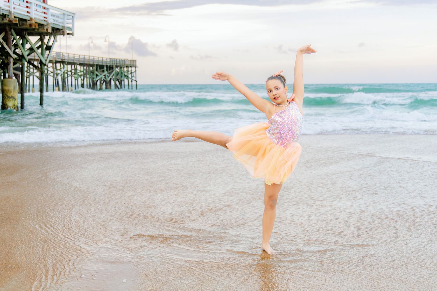 Young ballerina dancing gracefully on a beach near a pier, wearing a pink tutu, with ocean waves in the background.
