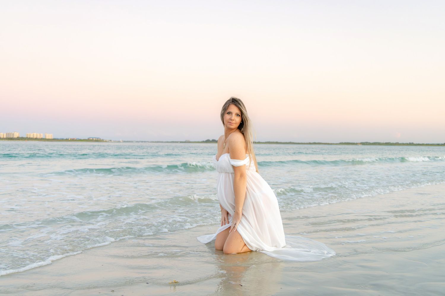 Pregnant woman in white off-shoulder dress posing in ocean waves at sunset beach.