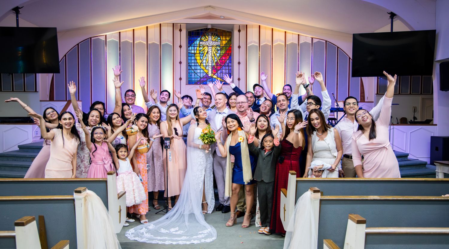 A joyful wedding party celebrates in a decorated church, with the bride and groom surrounded by guests in pink and formal attire.