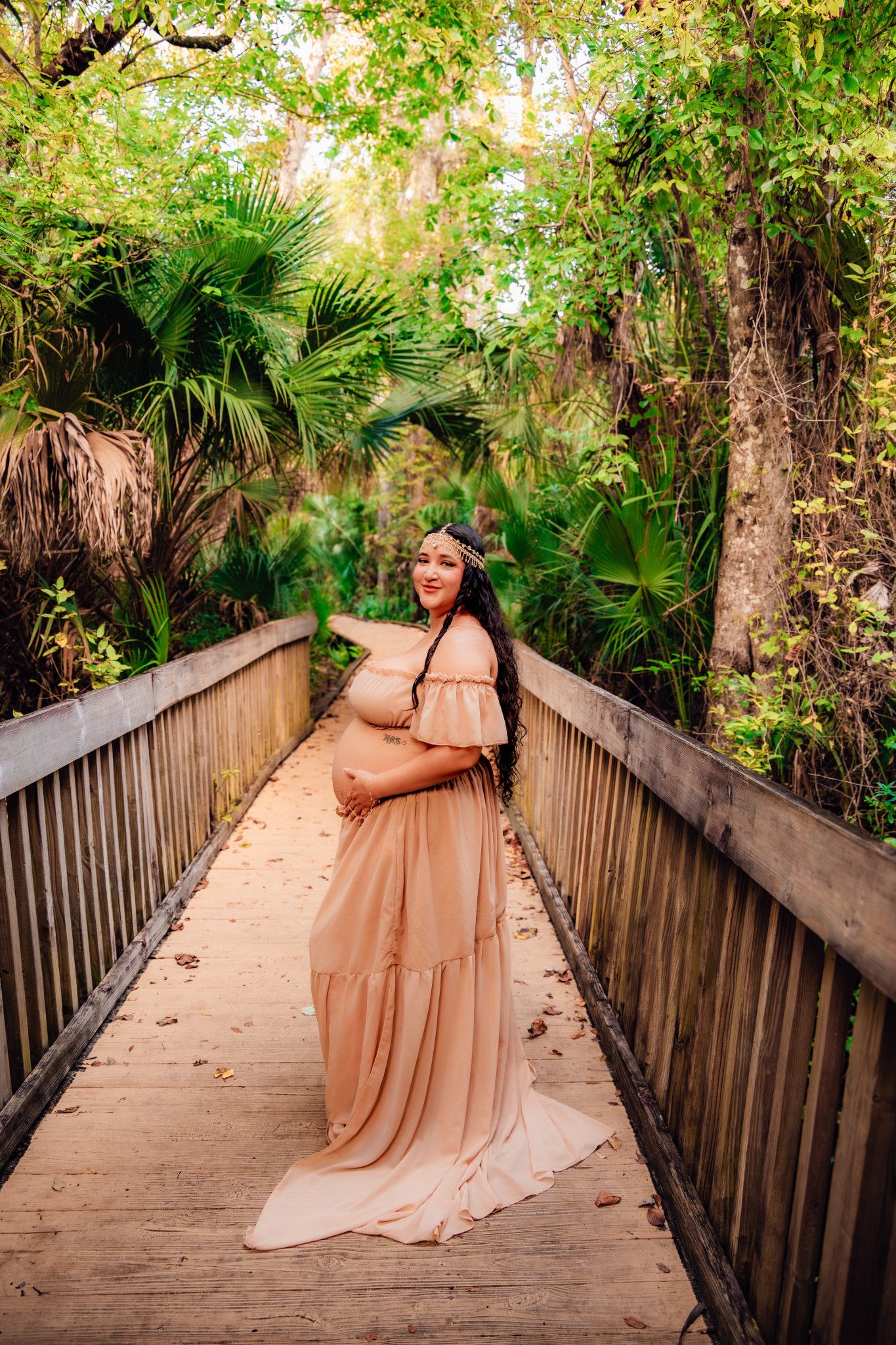 Pregnant woman in blush off-shoulder gown poses on wooden bridge surrounded by lush tropical greenery.