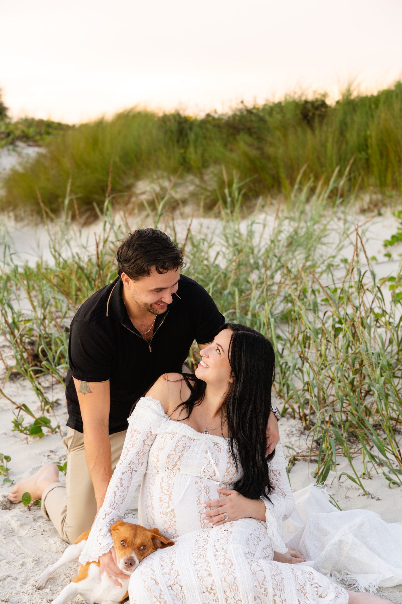 Pregnant couple sitting on beach sand dunes with dog, woman in white lace dress, man in black shirt, at sunset.