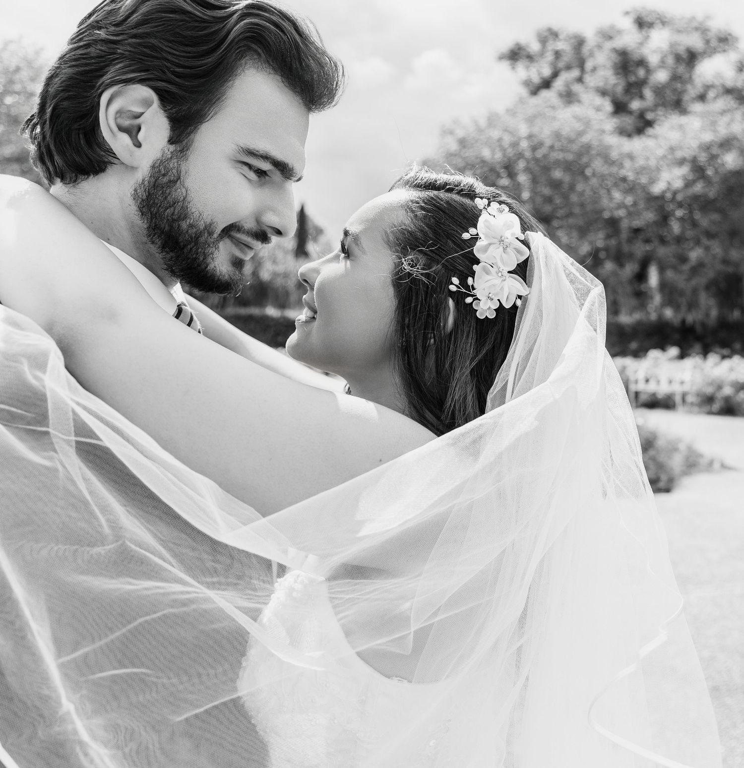 Black and white photo of a bride and groom sharing an intimate moment, wrapped in a flowing veil outdoors.