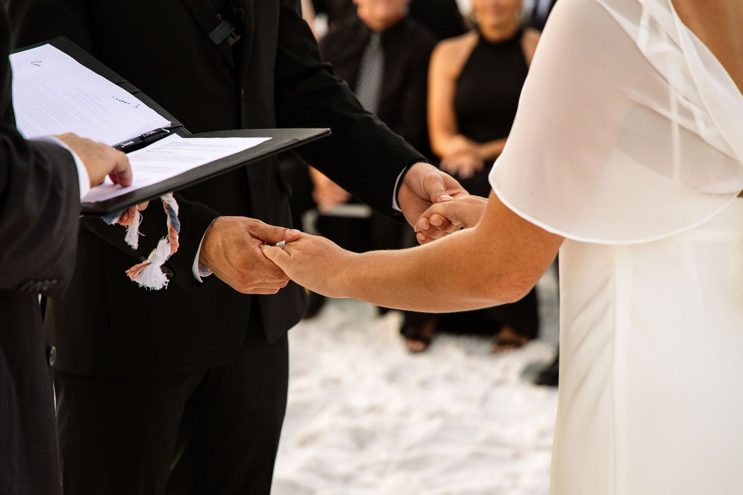 Groom placing wedding ring on bride's finger during ceremony, officiant holding book nearby.