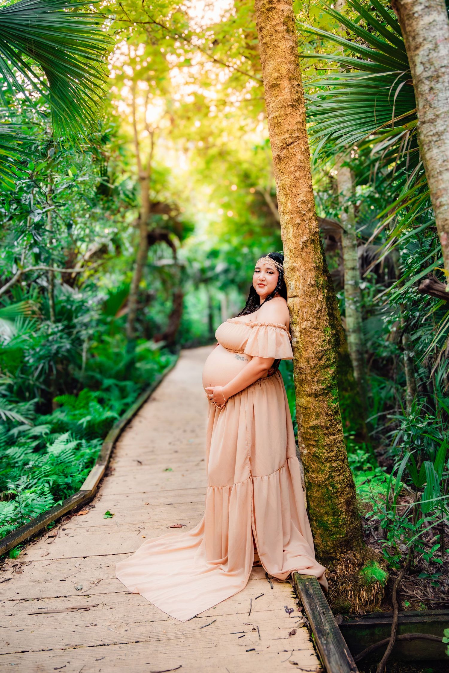 Pregnant woman in pink off-shoulder gown poses on tropical garden path surrounded by lush green palms.