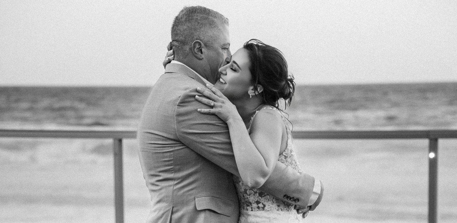 A romantic black and white photograph of a couple embracing and kissing on a beach boardwalk by the ocean.