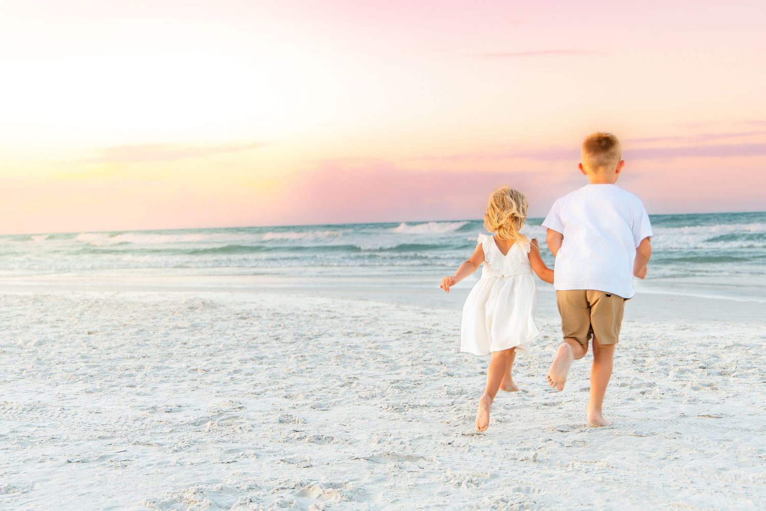 Two young children holding hands and running on a sandy beach at sunset in white and tan outfits.
