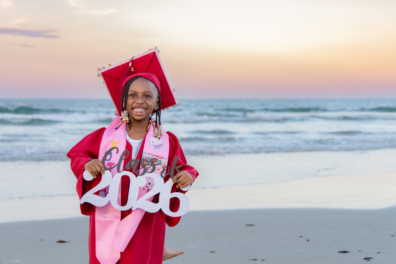 Young graduate in red cap and gown holds Class of 2026 sign on a beach at sunset.