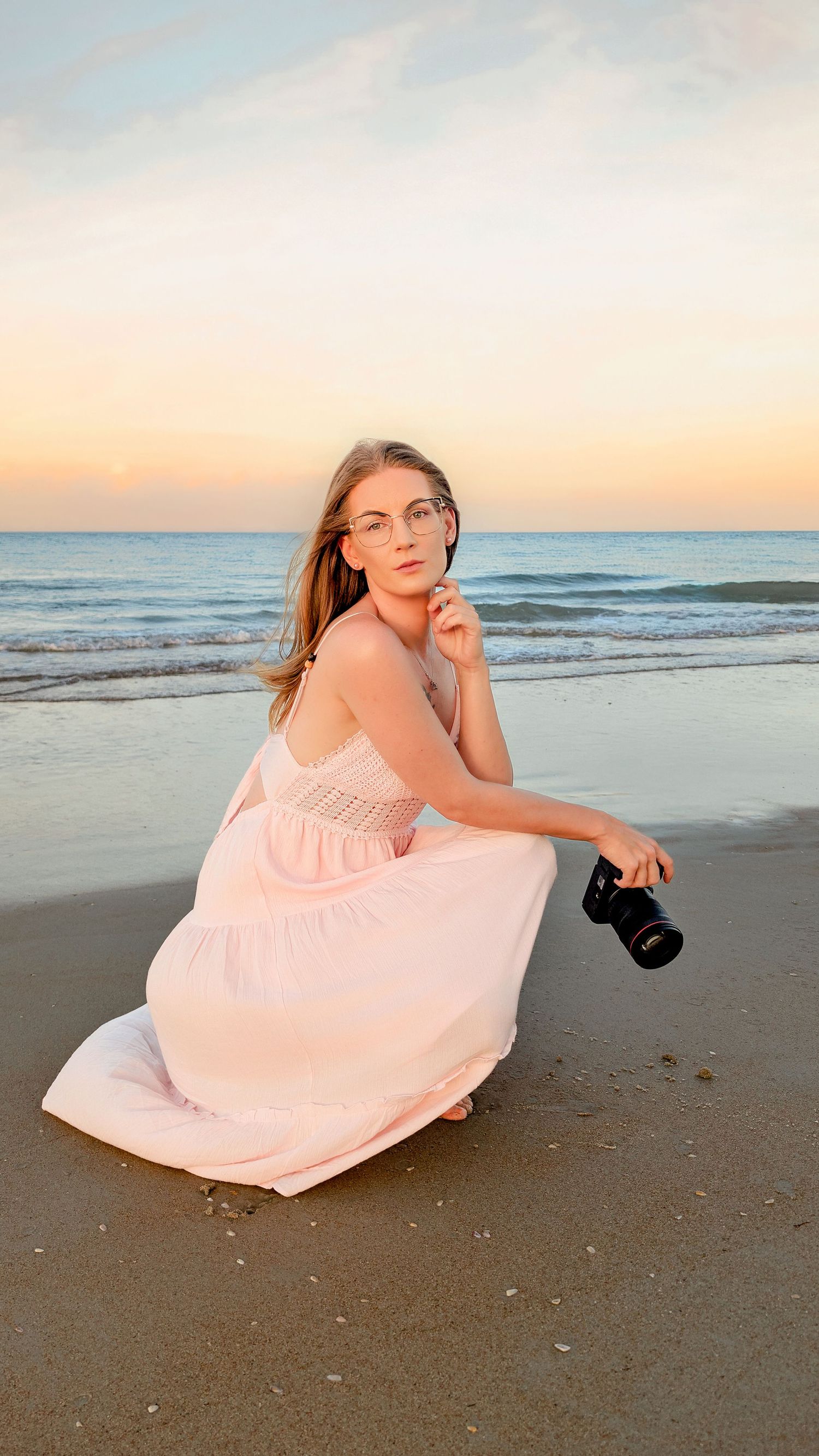 Young woman in pink dress holding camera poses on sandy beach at sunset with ocean waves in background.