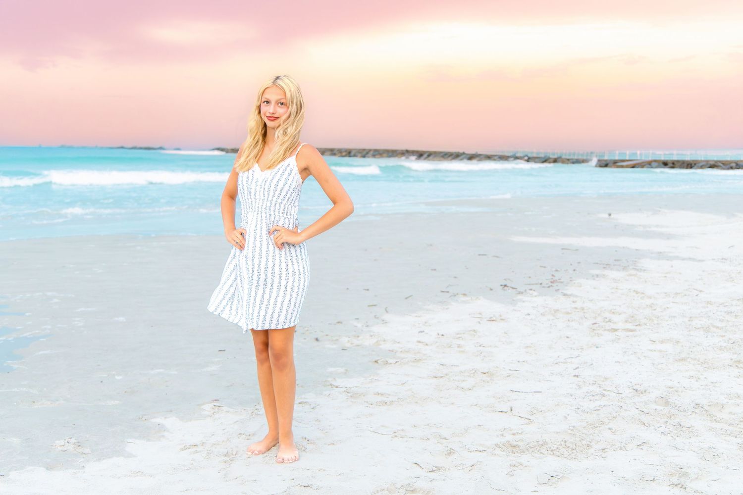 Blonde woman in striped white dress standing barefoot on a white sand beach at sunset.