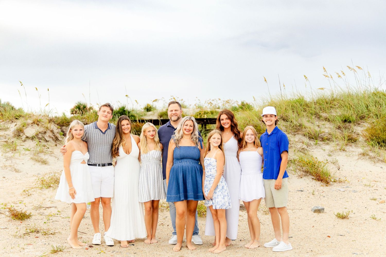 Large family group smiling together on a sandy beach with sea oats, dressed in coordinated blue and white outfits.