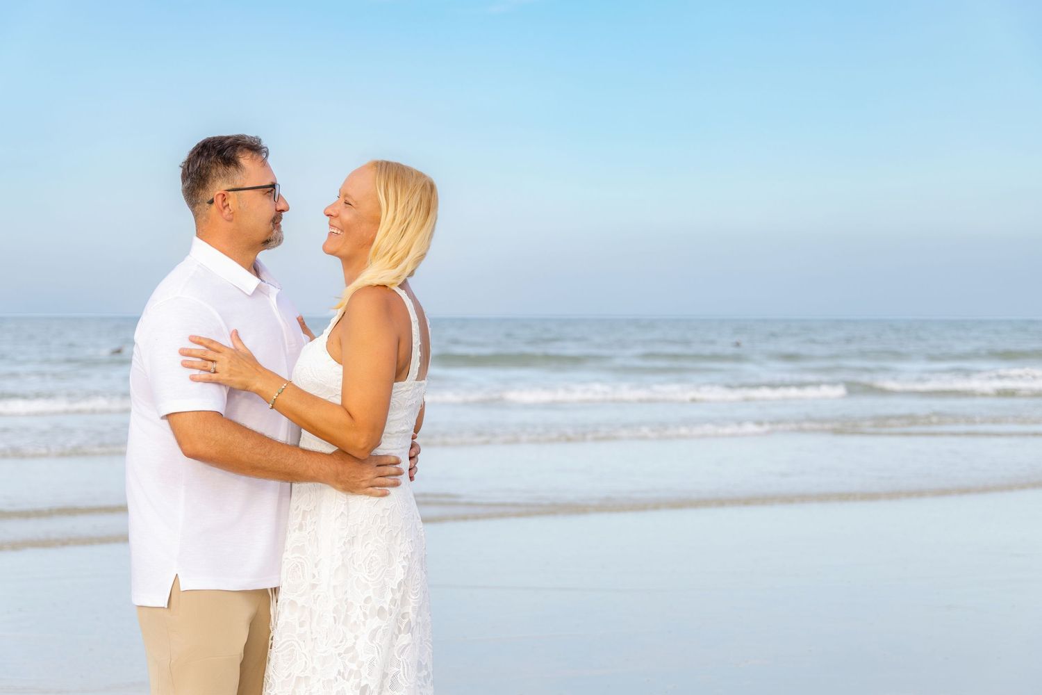 Couple embracing on a beach, woman in white lace dress, man in white shirt, with ocean waves in the background.