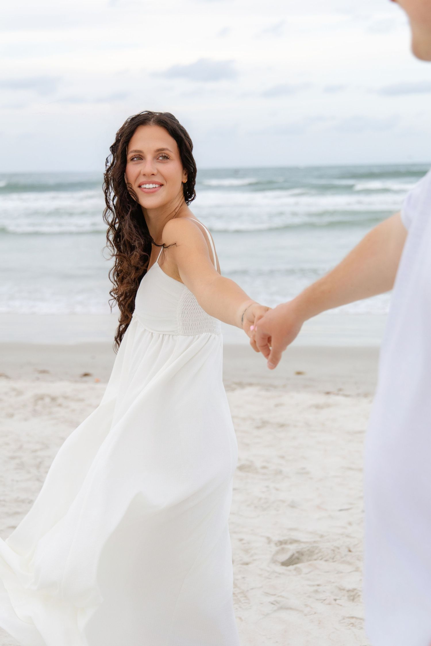 Smiling bride in white dress holds hands with groom on sandy beach with ocean waves in background.
