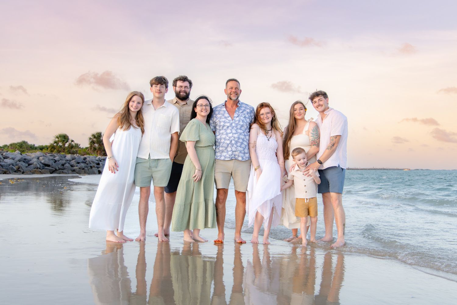 Large family group posing together on a tropical beach at sunset with pastel sky and ocean reflections.