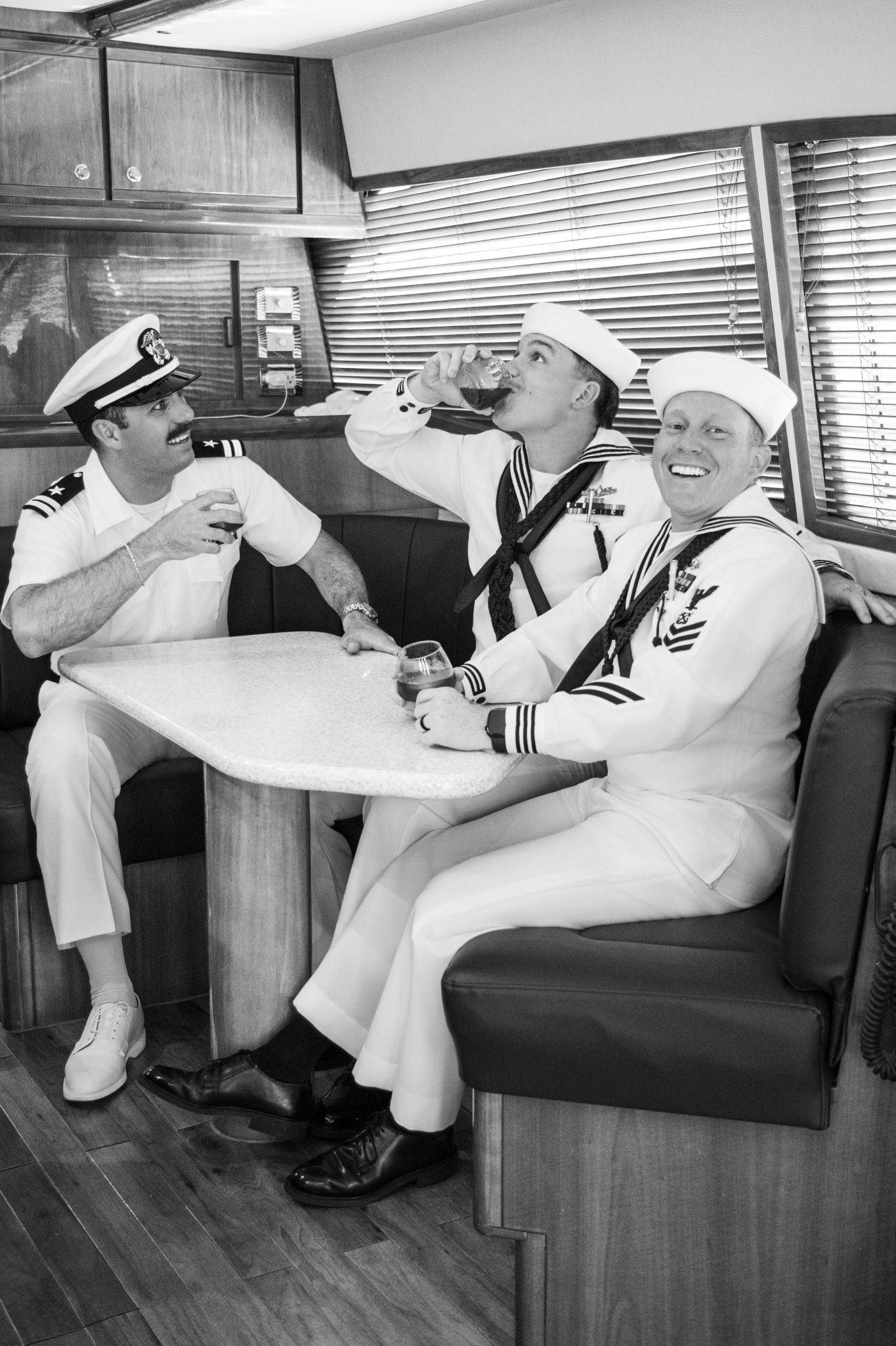 Three men in naval uniforms laugh and drink together at a booth table in a black and white photo.