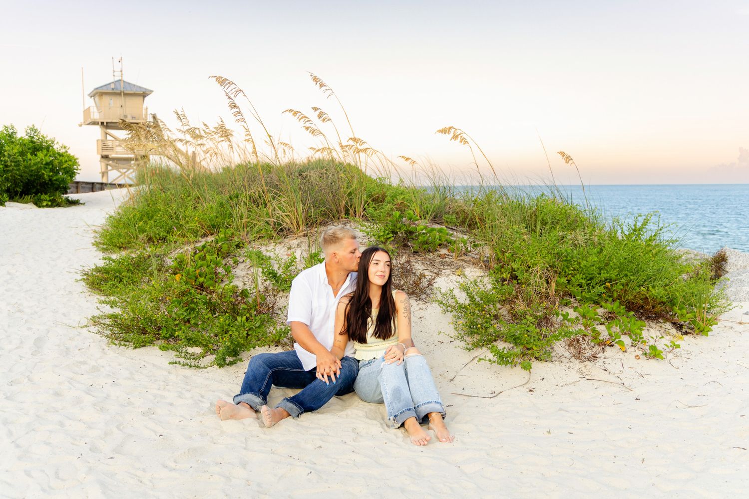 Couple sitting together on white sand beach at sunset with sea oats and a lifeguard tower in the background.