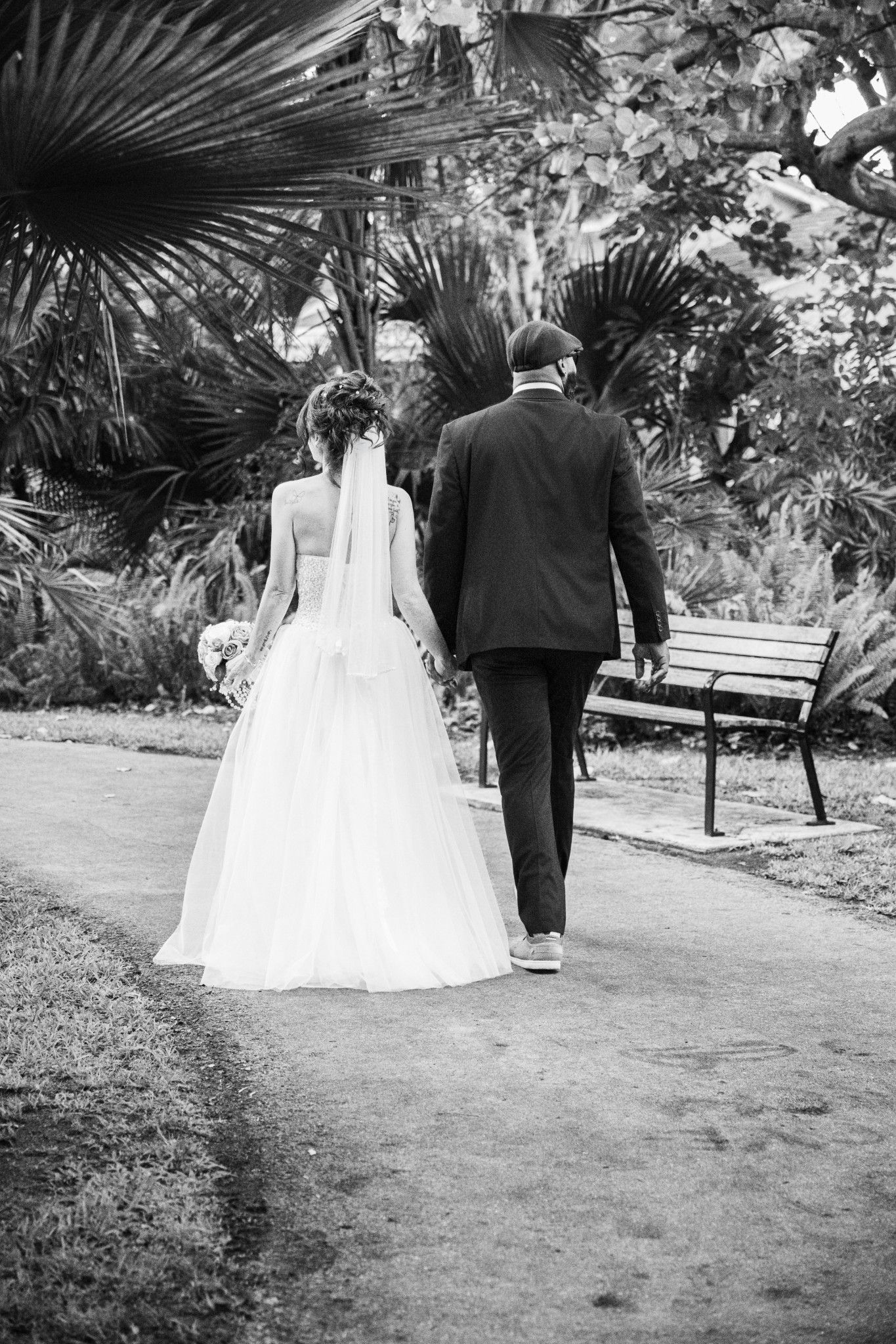 A couple in wedding attire walks along a garden path surrounded by palm trees and tropical foliage in black and white.