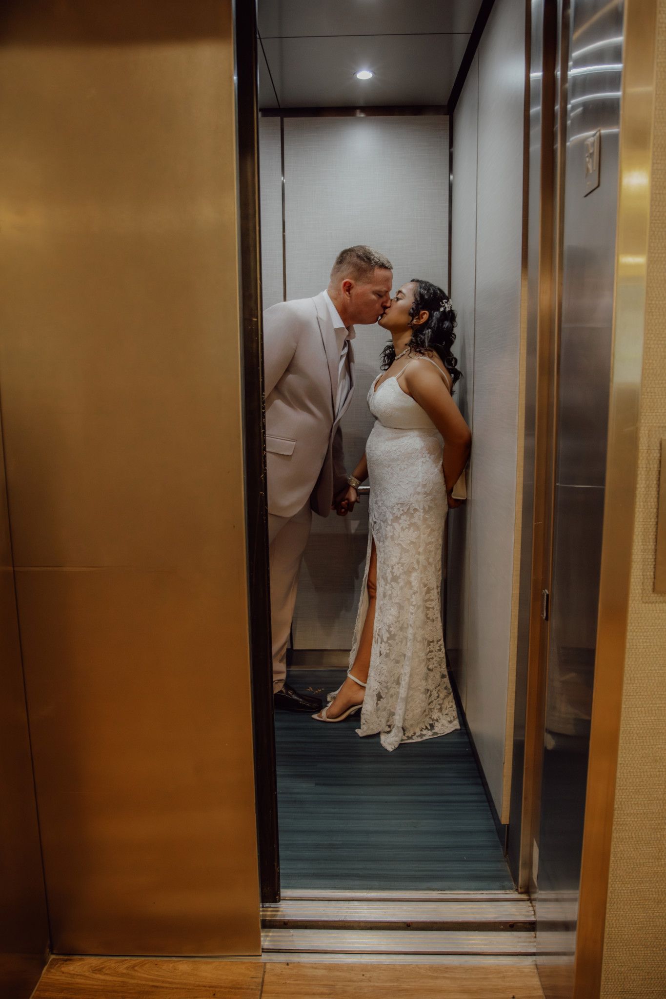 Bride and groom sharing a romantic kiss inside a golden elevator, bride in lace gown, groom in white suit.