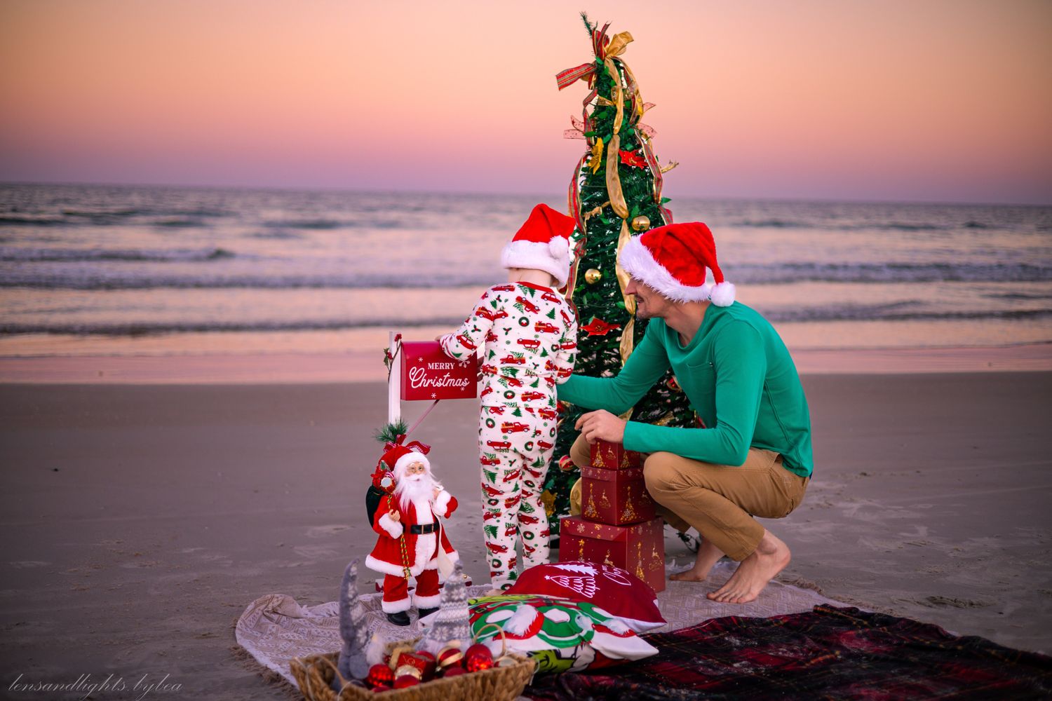 A festive Christmas scene on the beach at sunset with a decorated tree, presents, and Santa decorations in the sand.
