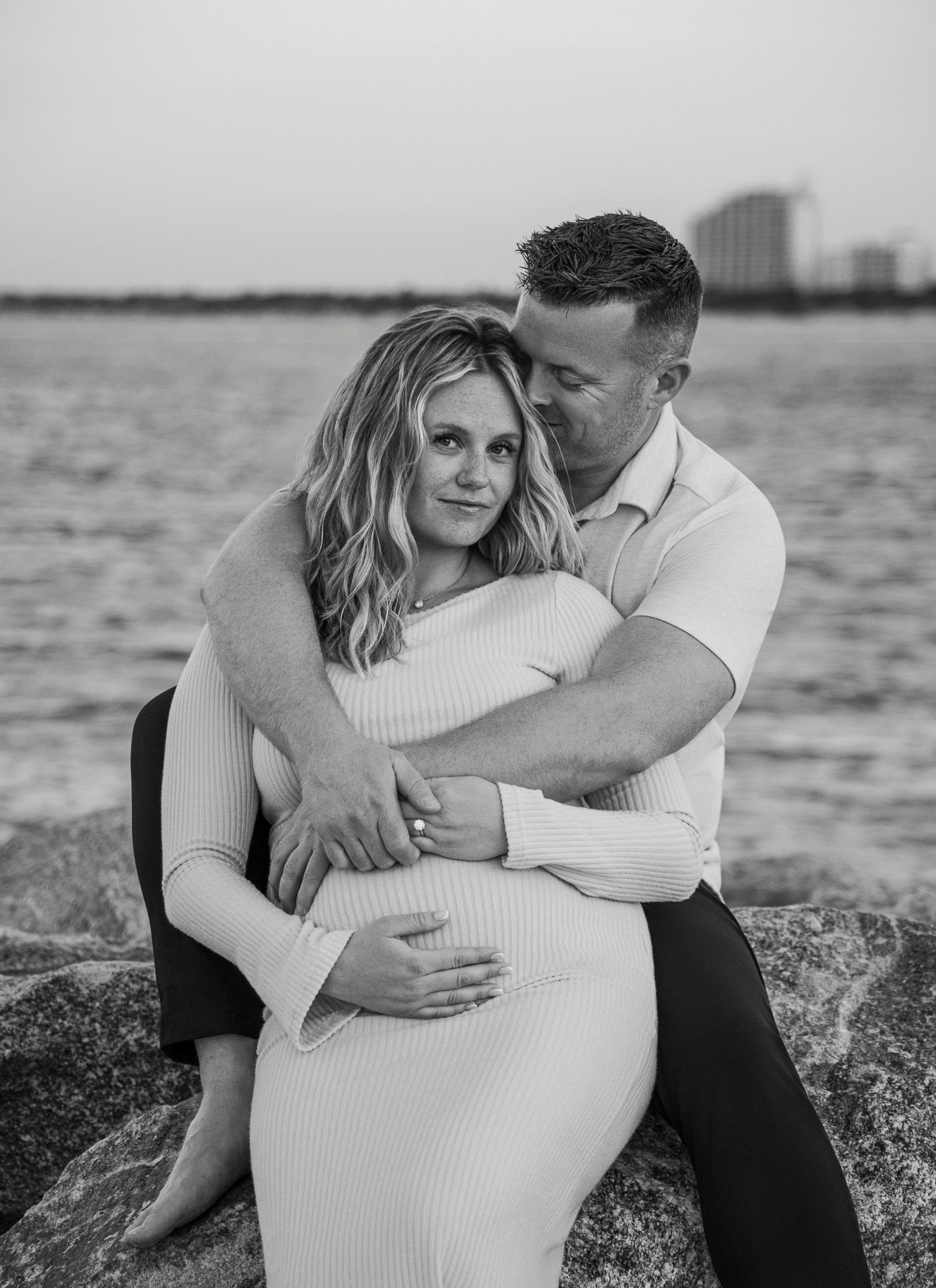 A couple embraces intimately on a rocky beach during a romantic black and white maternity photoshoot at sunset.