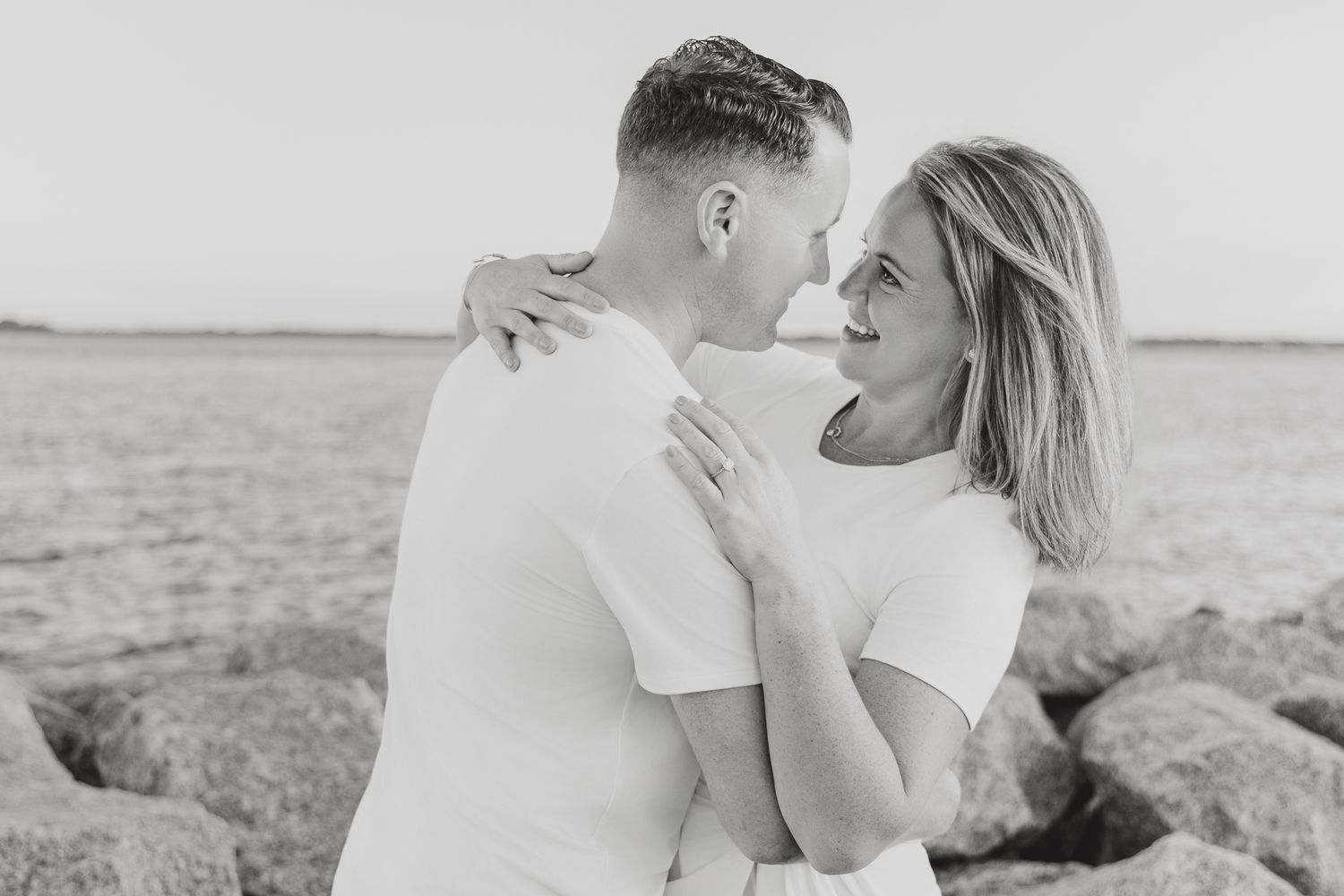 Black and white photo of a couple embracing and smiling at each other on a rocky beach.