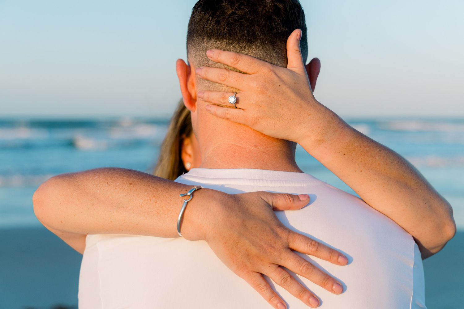 Couple embracing on a beach at sunset, woman wearing an engagement ring and bracelet hugging man in white shirt.