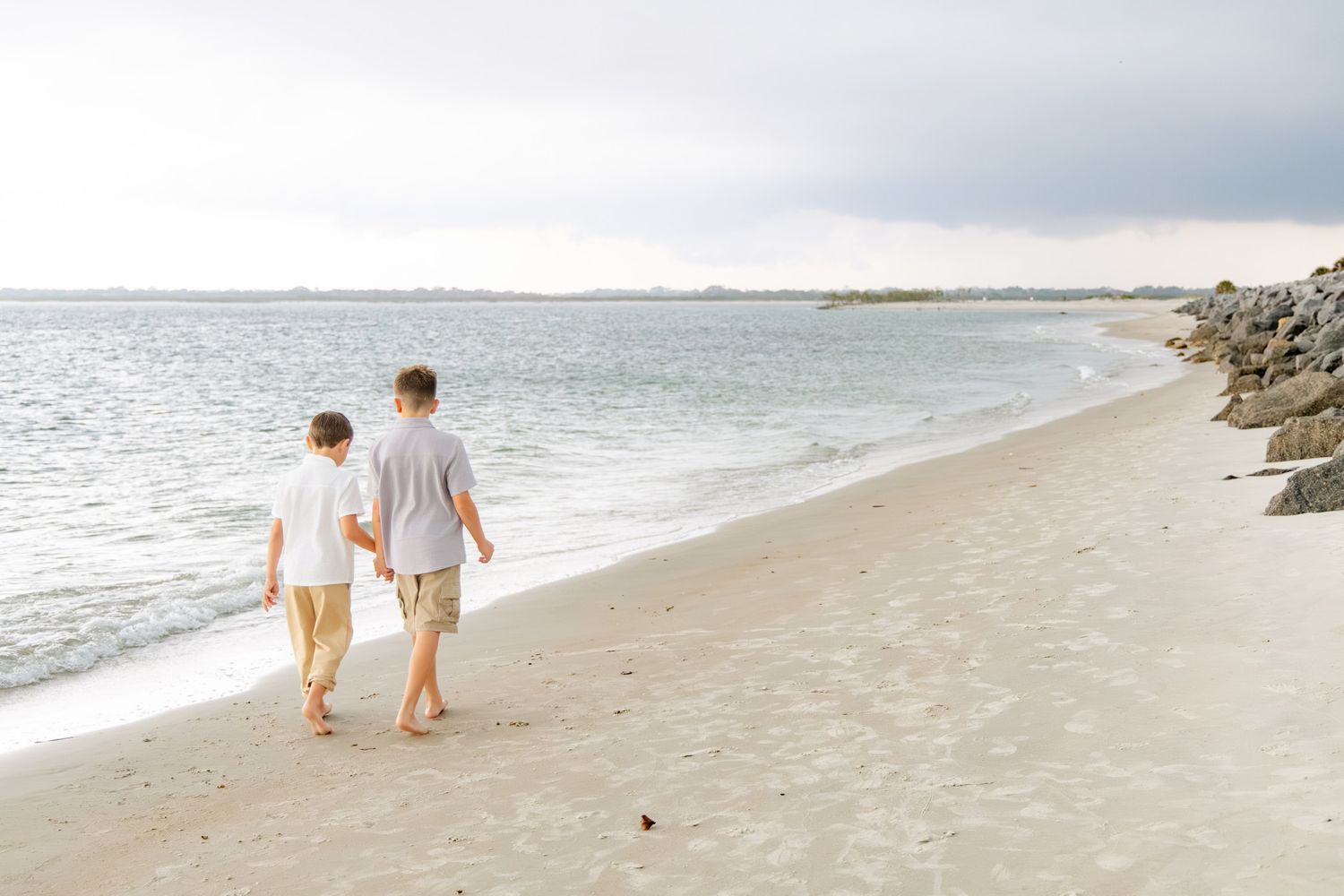 Two boys walking hand in hand along a sandy beach shoreline with gentle waves and rocky jetty in background.