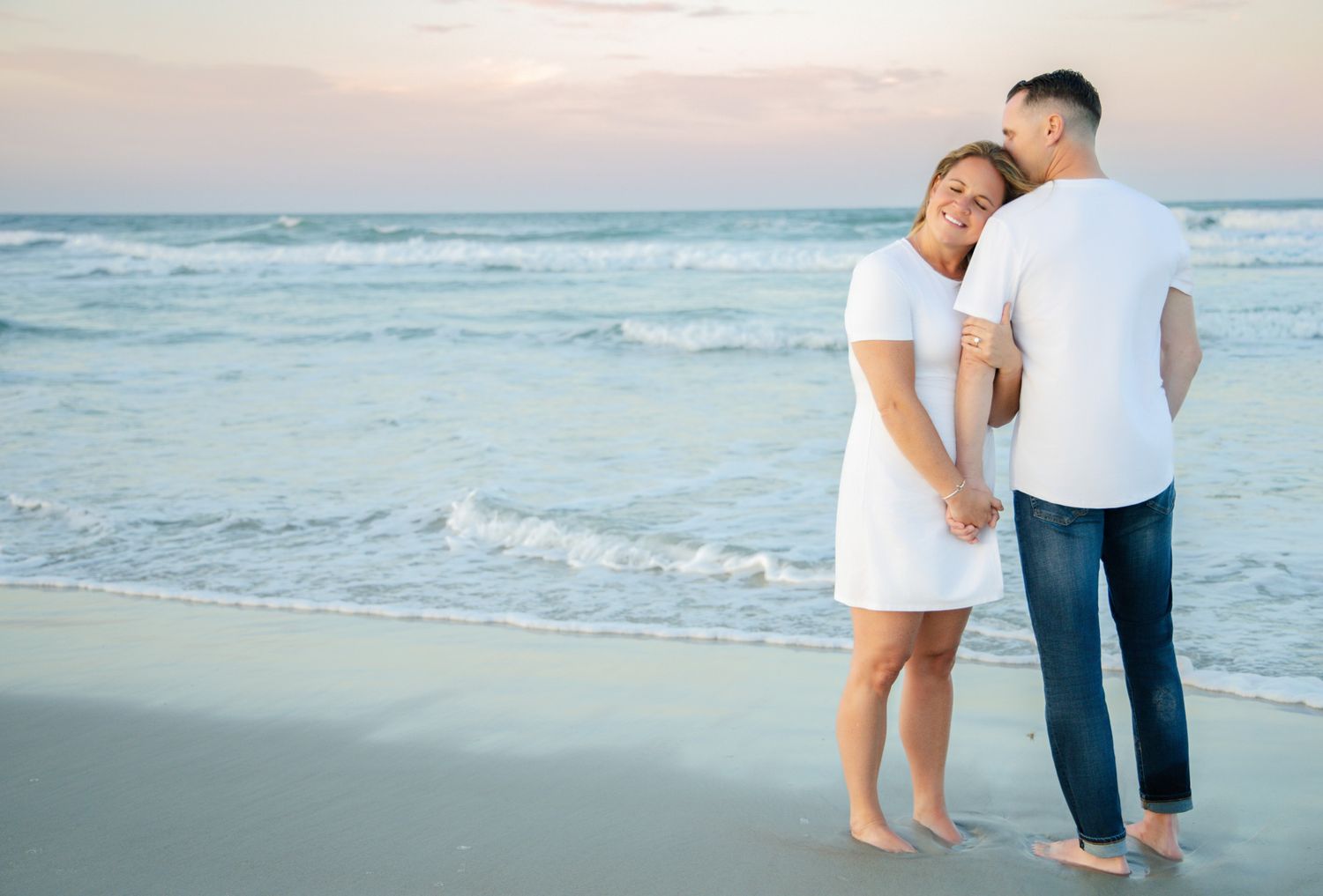 Couple embracing on a sandy beach at sunset, wearing white outfits, with gentle ocean waves in the background.