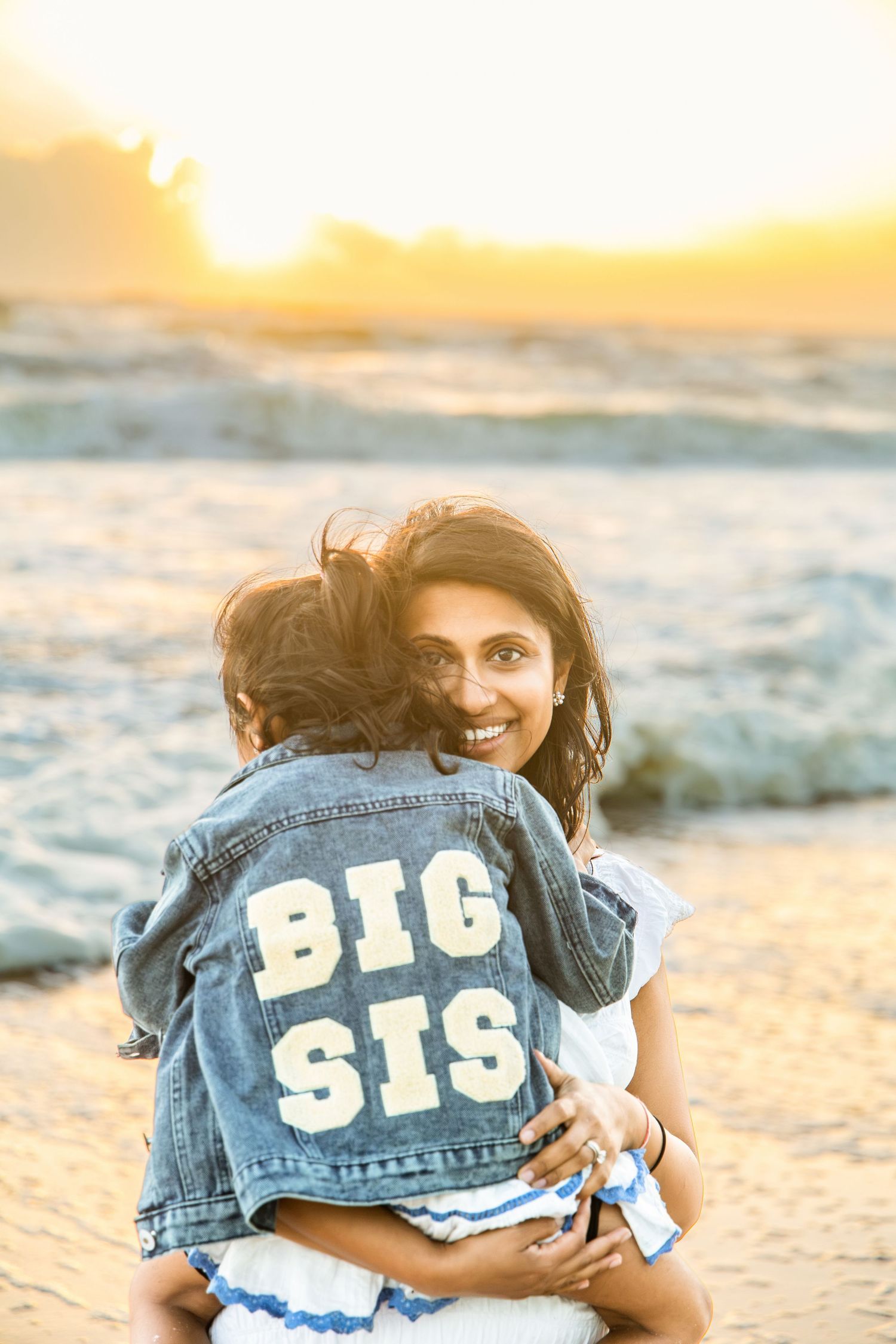 Smiling woman hugs a child in a denim jacket reading BIG SIS on a sunset beach with ocean waves.