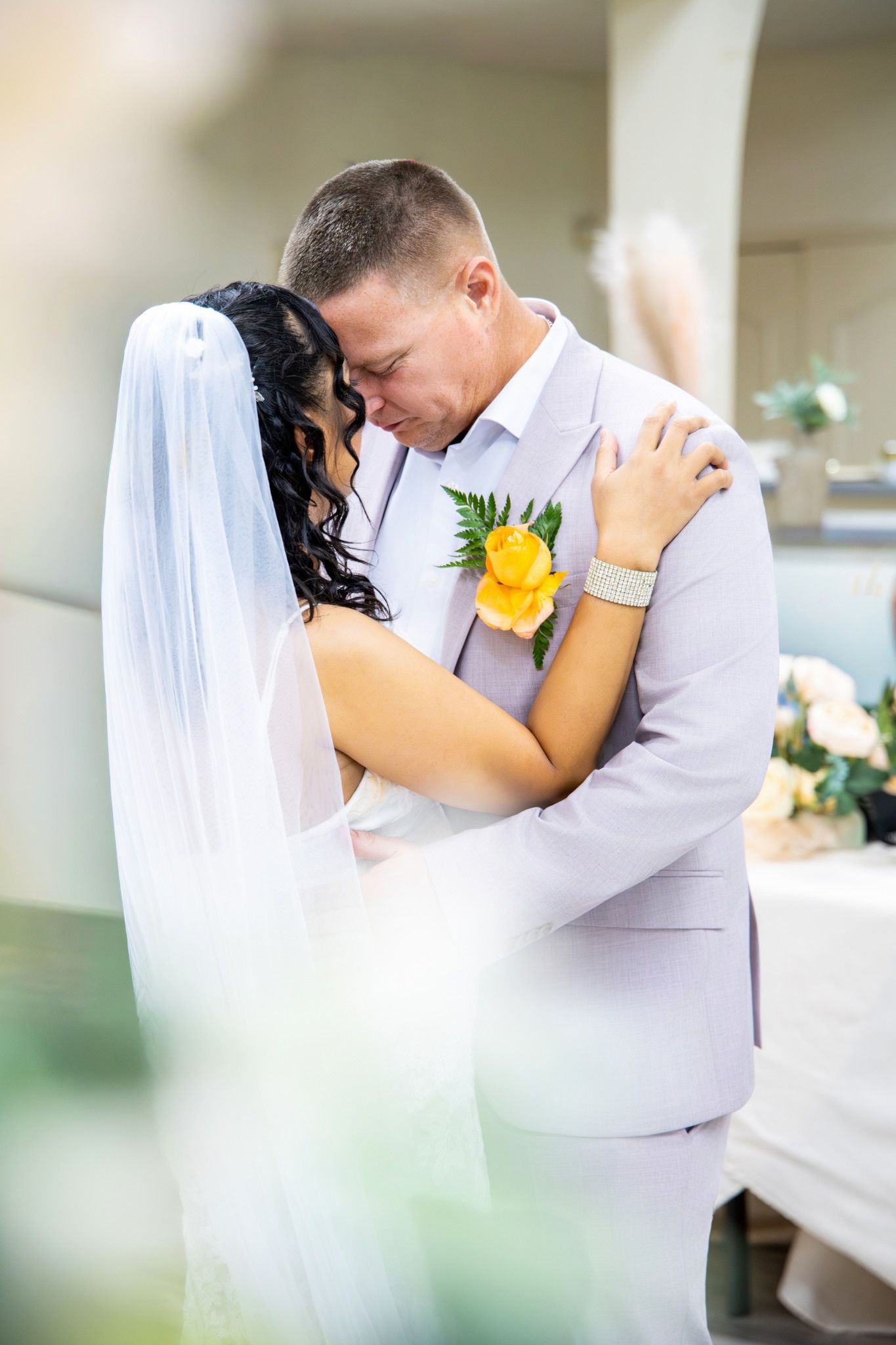 Bride and groom share an intimate embrace during wedding ceremony, bride wearing veil, groom in light gray suit with yellow boutonniere.
