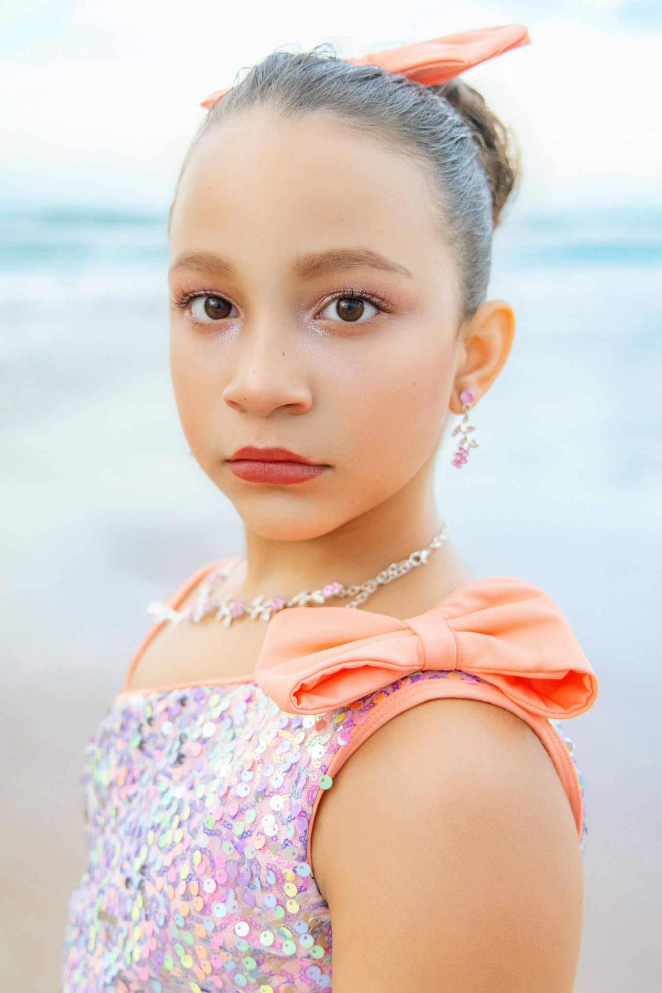 Young girl in sequin dress with coral bow, wearing jewelry, poses on beach with ocean backdrop.