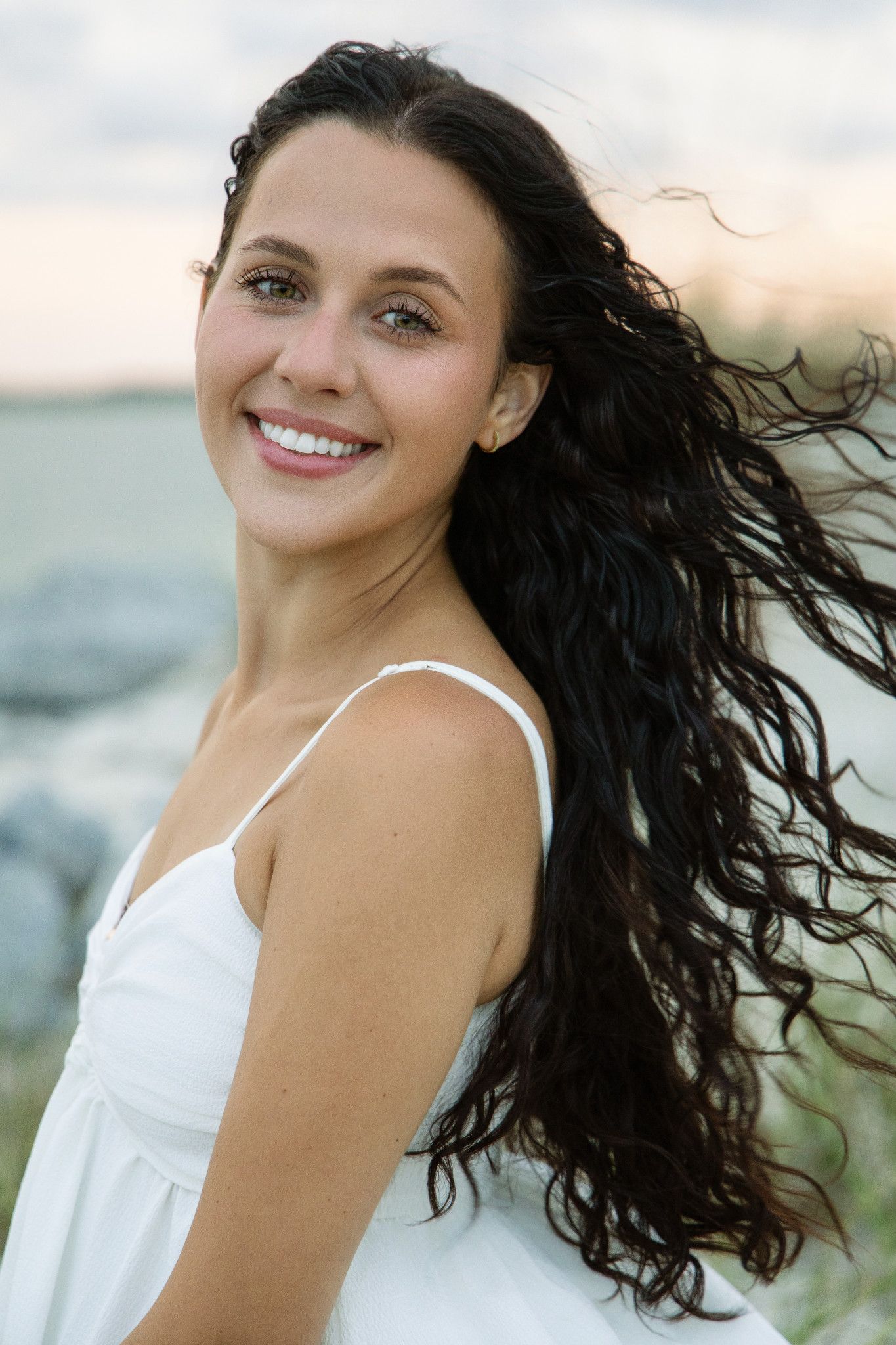 Smiling young woman with long dark curly hair wearing a white sundress outdoors near the water at sunset.